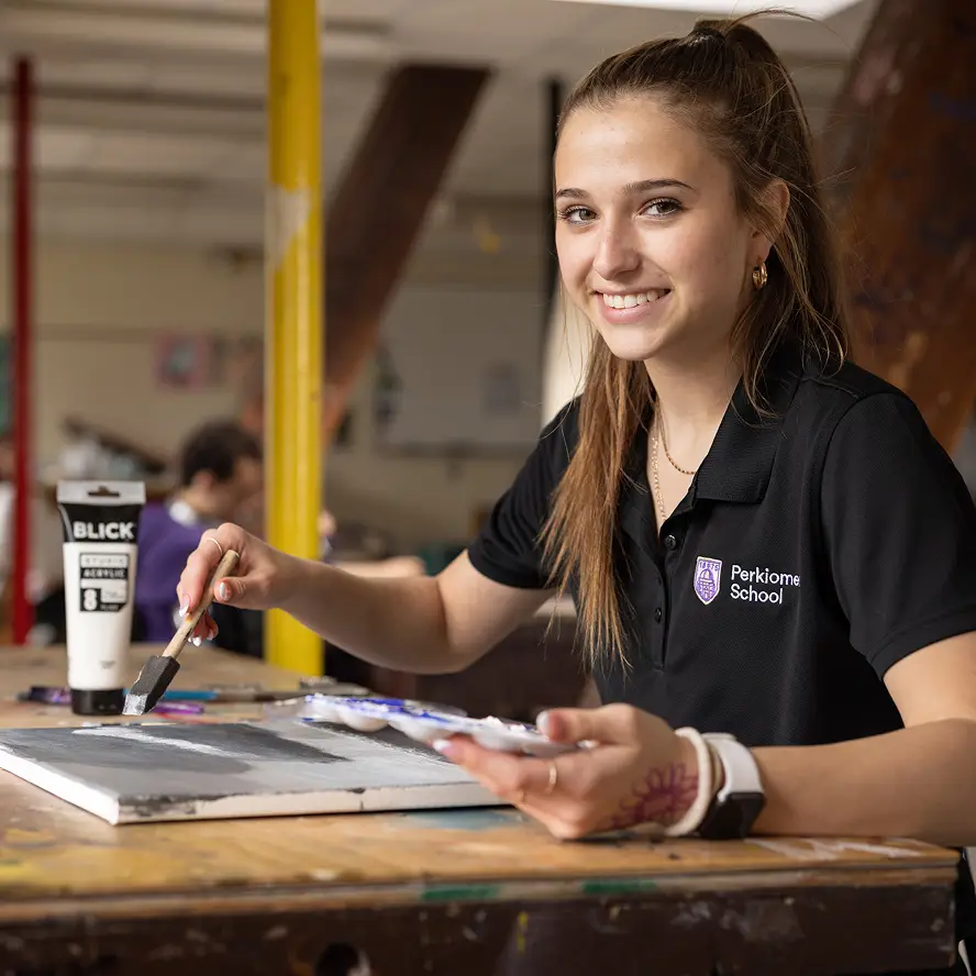 Smiling student in a Perkiome School polo shirt painting on a canvas at a wooden table.