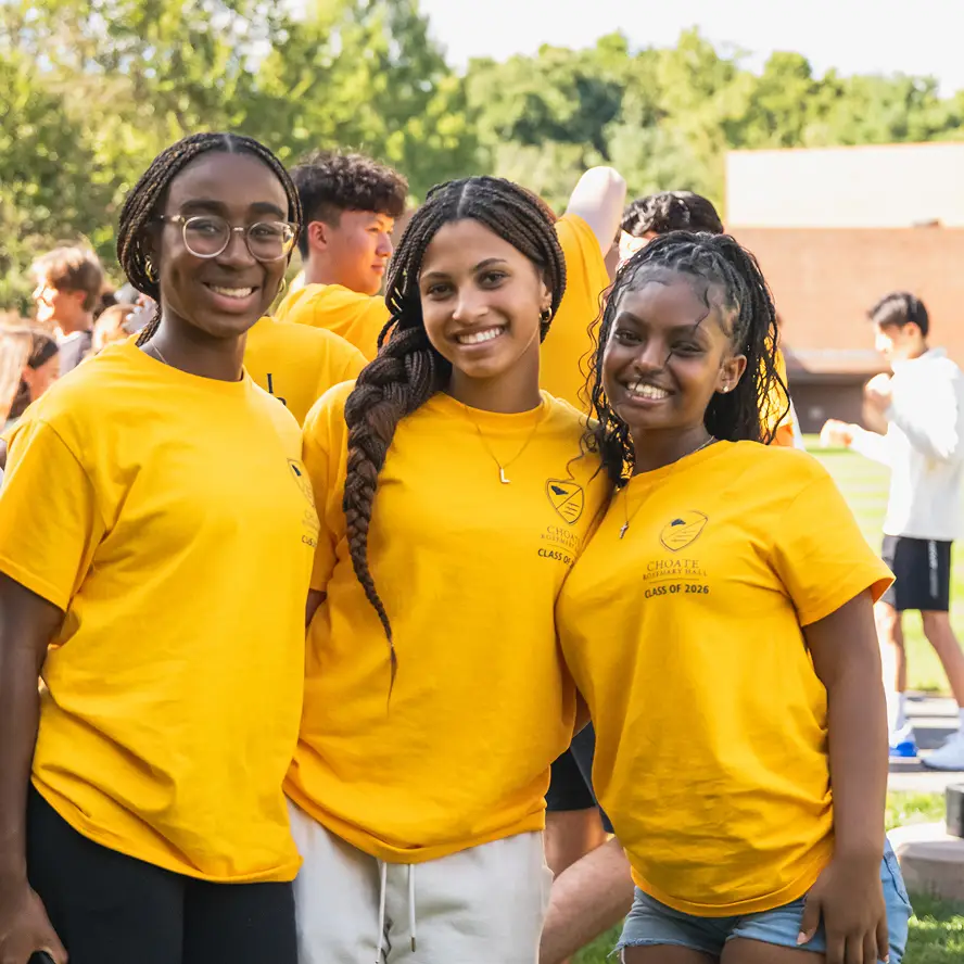 Three smiling young women in yellow Class of 2026 t-shirts standing outdoors with trees blurred in the background.