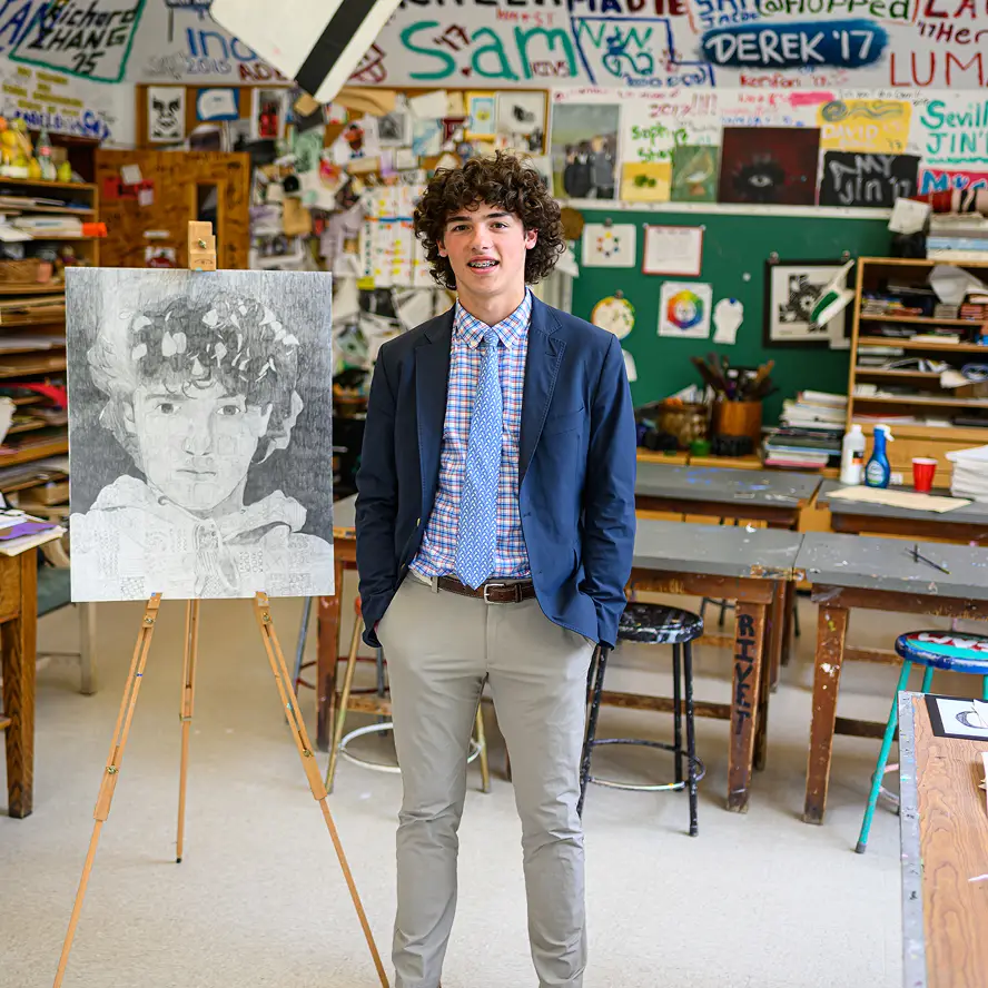 Young man with curly hair wearing a blue blazer, checkered shirt, and tie stands next to an easel displaying a pencil drawing of his portrait in an art classroom.