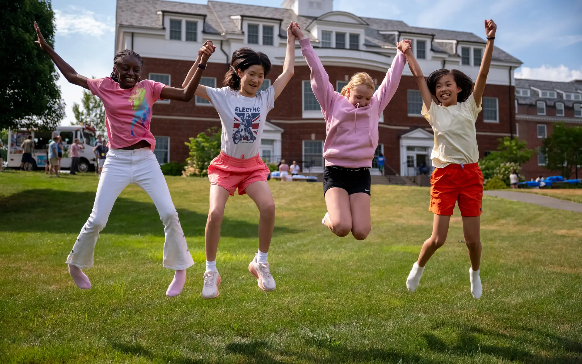Four diverse children holding hands and joyfully jumping on green grass in front of a brick building on a sunny day.
