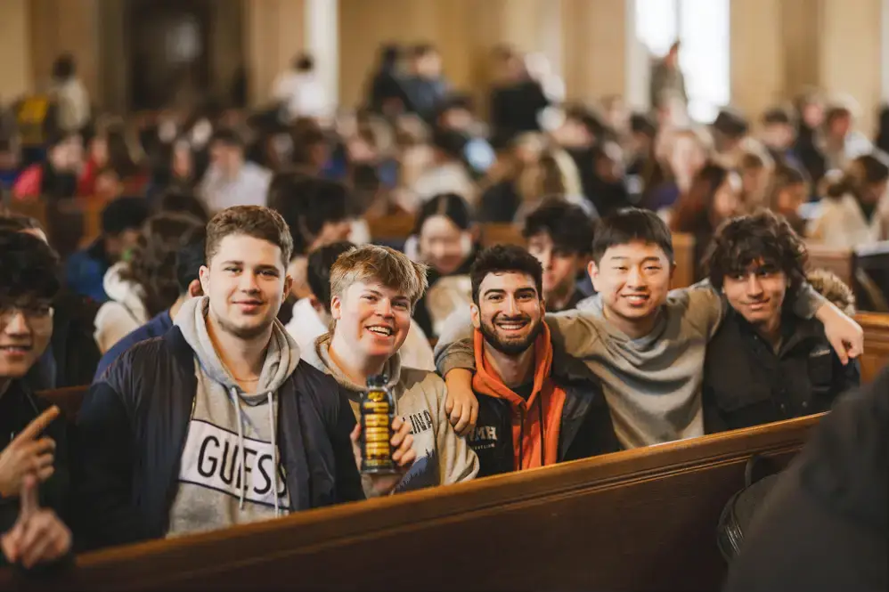 A group of five diverse young men sitting closely together on wooden benches in a crowded indoor setting, smiling and posing for the photo.