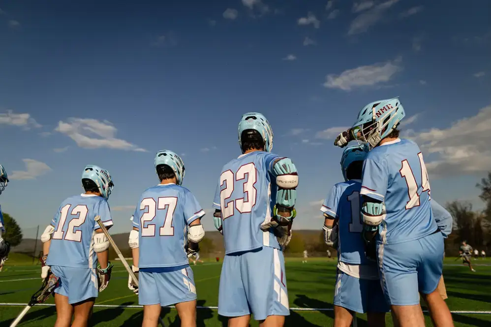Lacrosse players in light blue uniforms and helmets standing together on a green field under a blue sky.