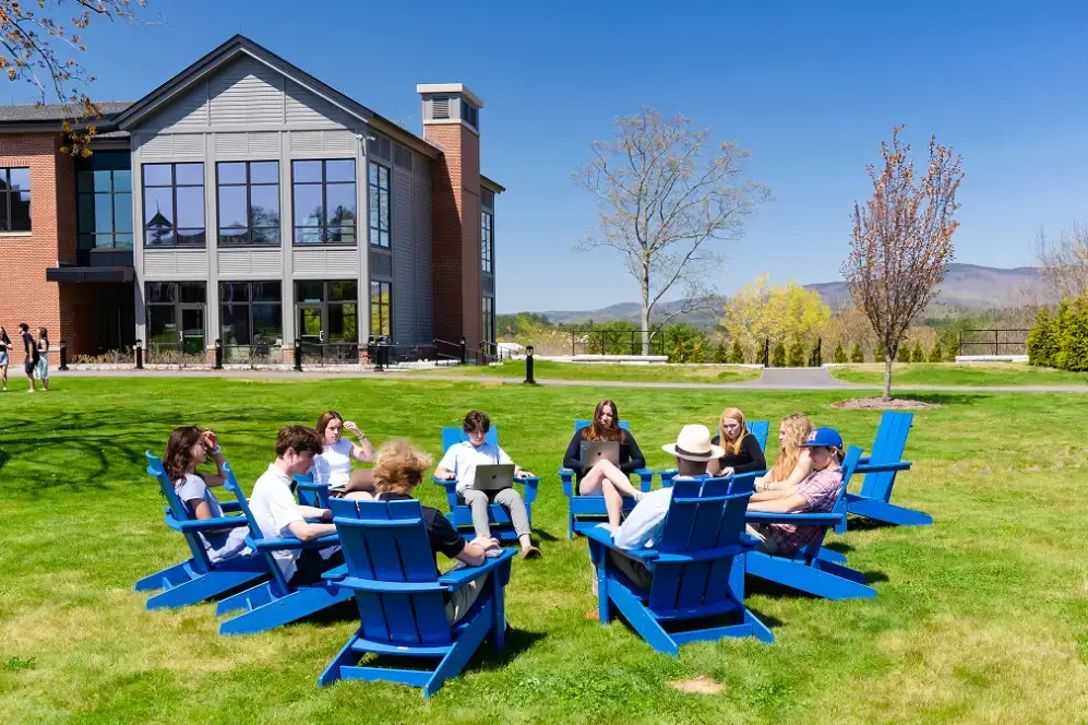 Group of young people sitting and working on laptops in blue Adirondack chairs arranged in a circle on a grassy lawn outside a modern building under a clear blue sky.