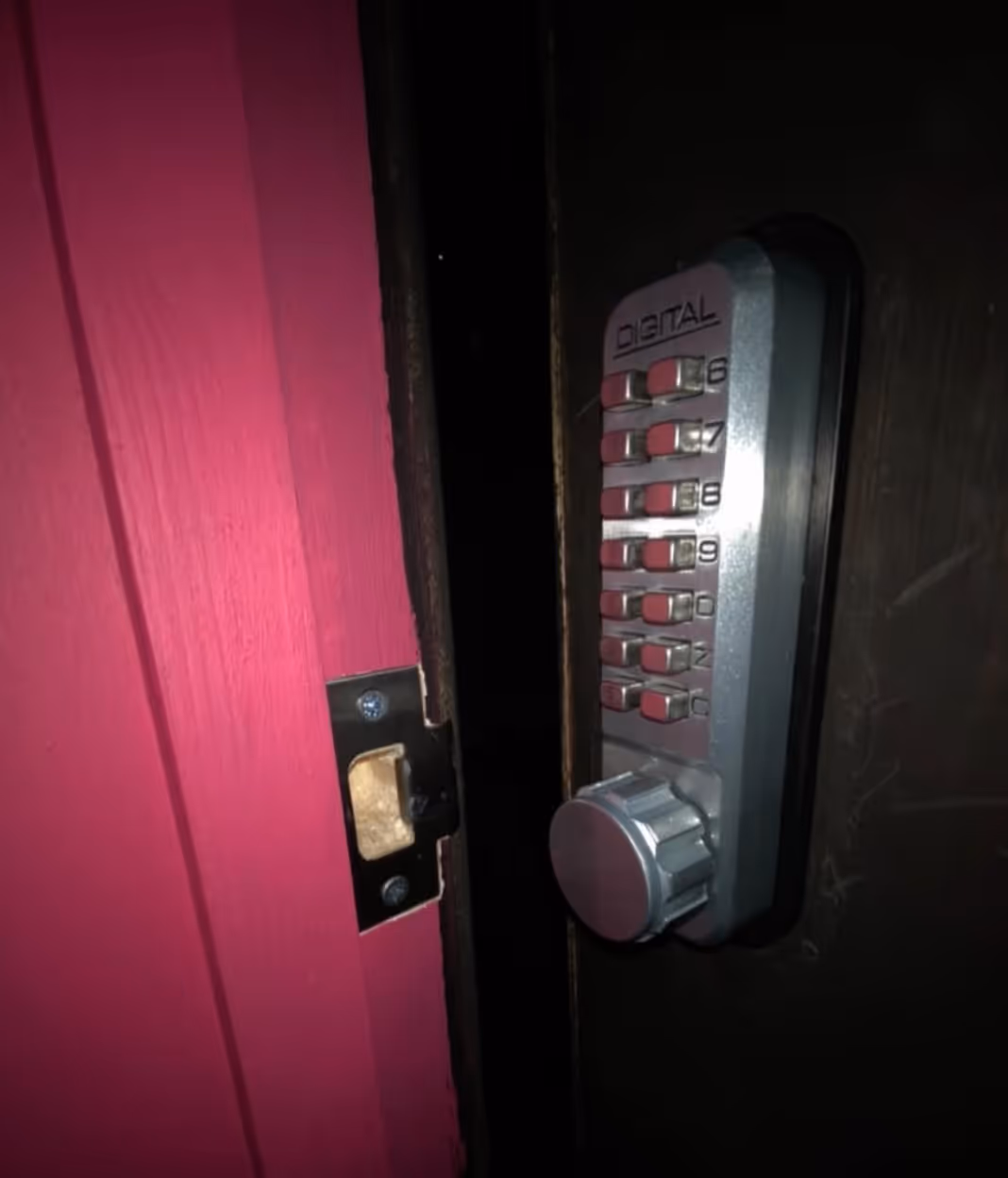 Close-up of a digital keypad lock with a silver dial on a dark door next to a pink door frame.