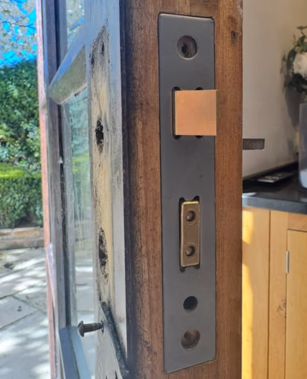 Close-up of a wooden door edge showing a metal latch bolt and part of the door lock mechanism with screws and a glass panel window.
