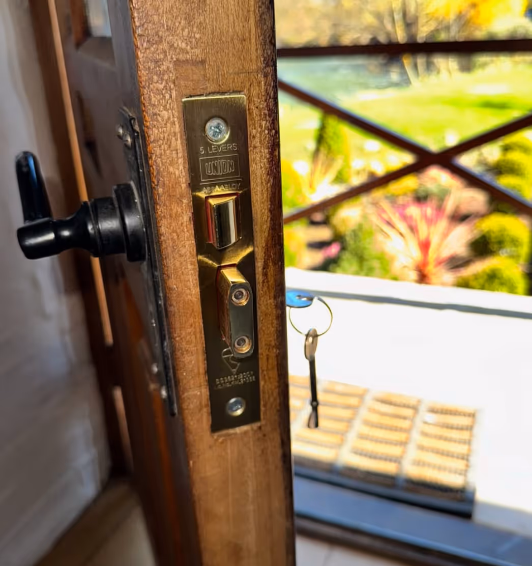 Close-up of a wooden door's lock mechanism with a key inserted, showing a garden outside through the open door.