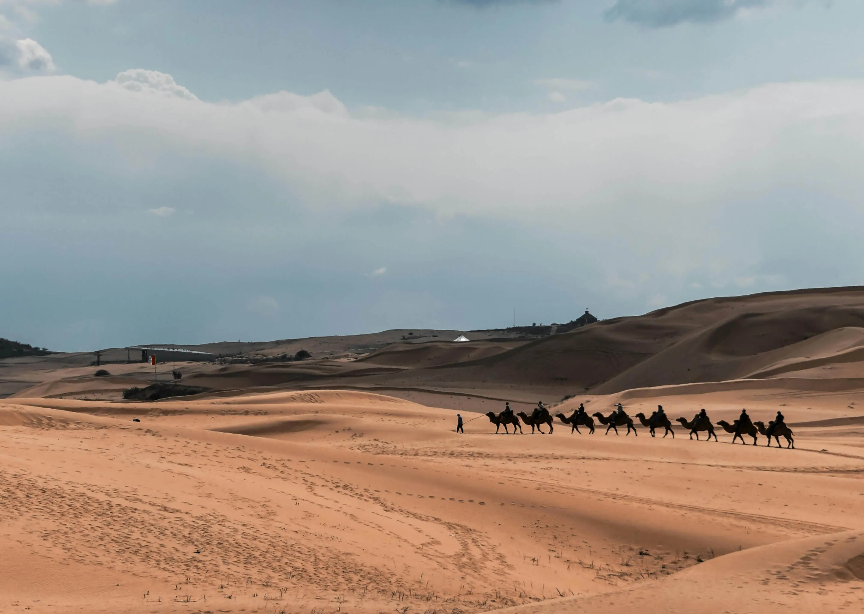 Line of people riding camels across sunlit sand dunes under a cloudy sky.