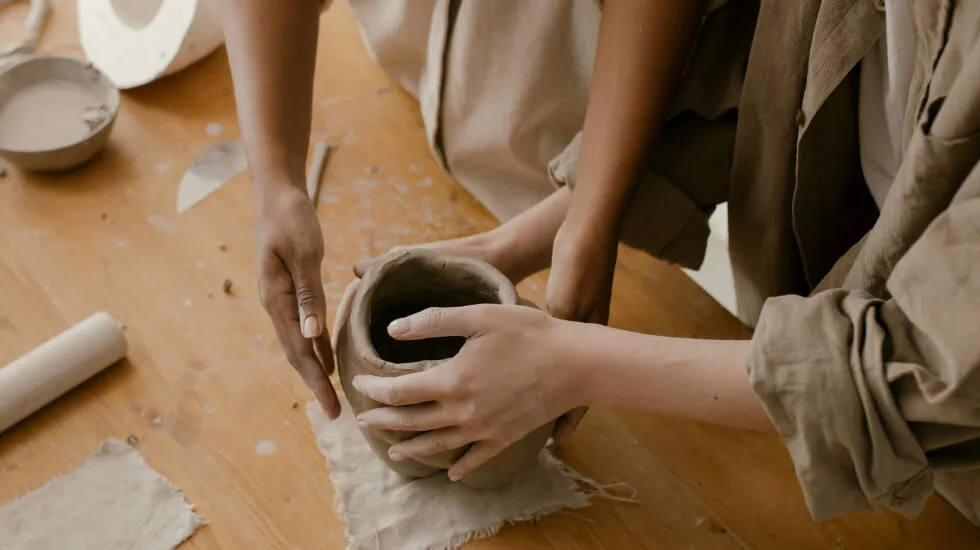 Two people shaping a clay pot together on a wooden table with pottery tools nearby.