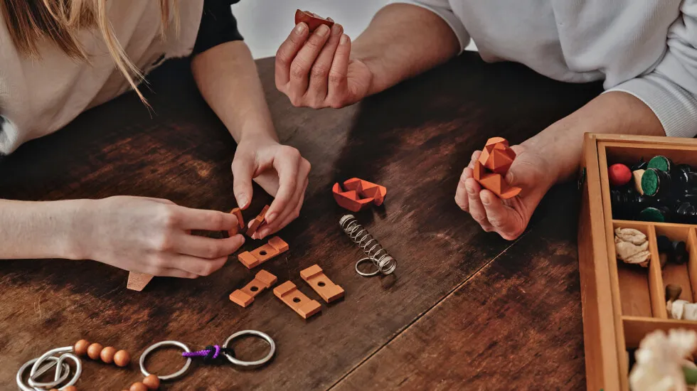 Two people assembling wooden interlocking puzzle pieces on a wooden table with additional puzzle parts and a box of game pieces nearby.