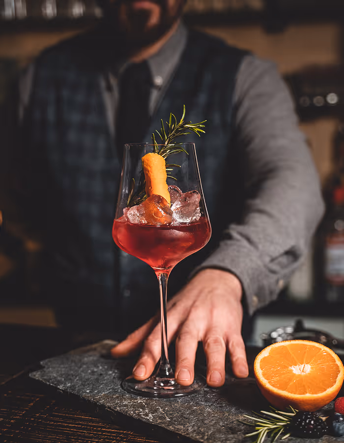 Ein Barkeeper präsentiert ein rotes Cocktailglas mit Eiswürfeln, einem Zweig Rosmarin und einer Orangenschale.