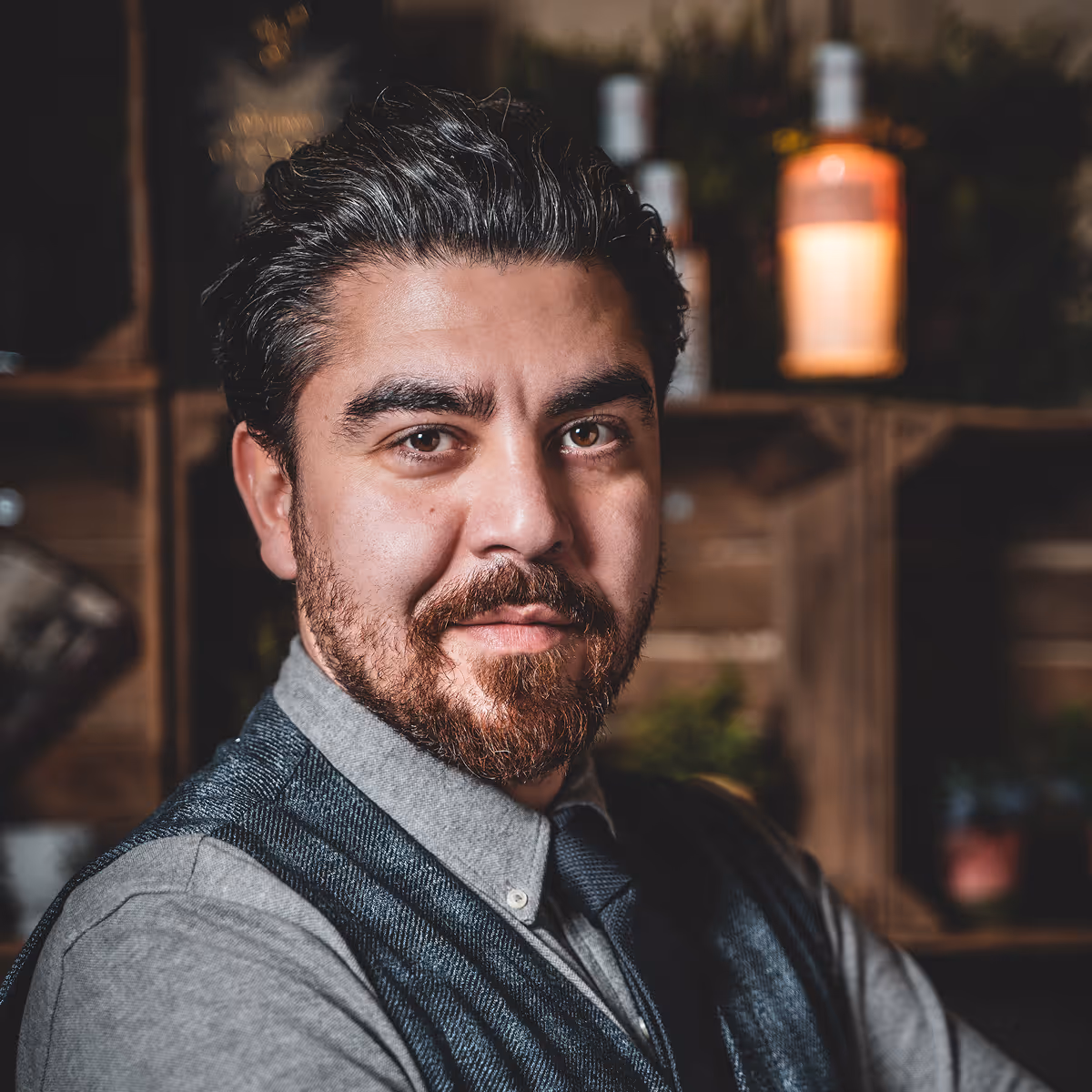 Mann mit Bart und dunklen Haaren lächelt in einer Bar oder einem Café mit Regalen und Flaschen im Hintergrund.