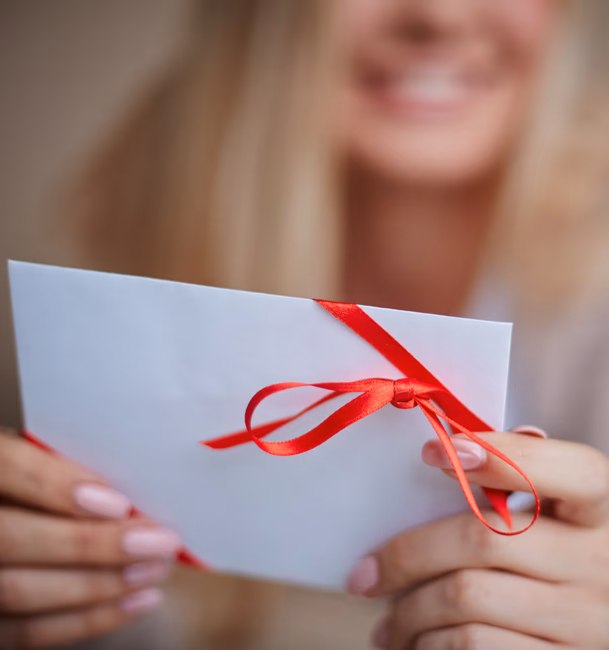 Frau hält einen weißen Briefumschlag mit rotem Schleifenband und lächelt im Hintergrund.