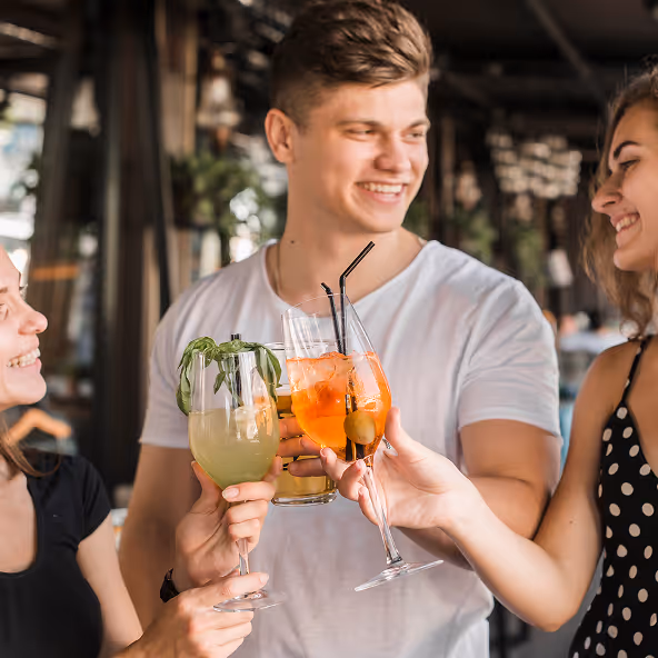 Drei junge Erwachsene stoßen mit Cocktails in Gläsern an und lächeln sich an.