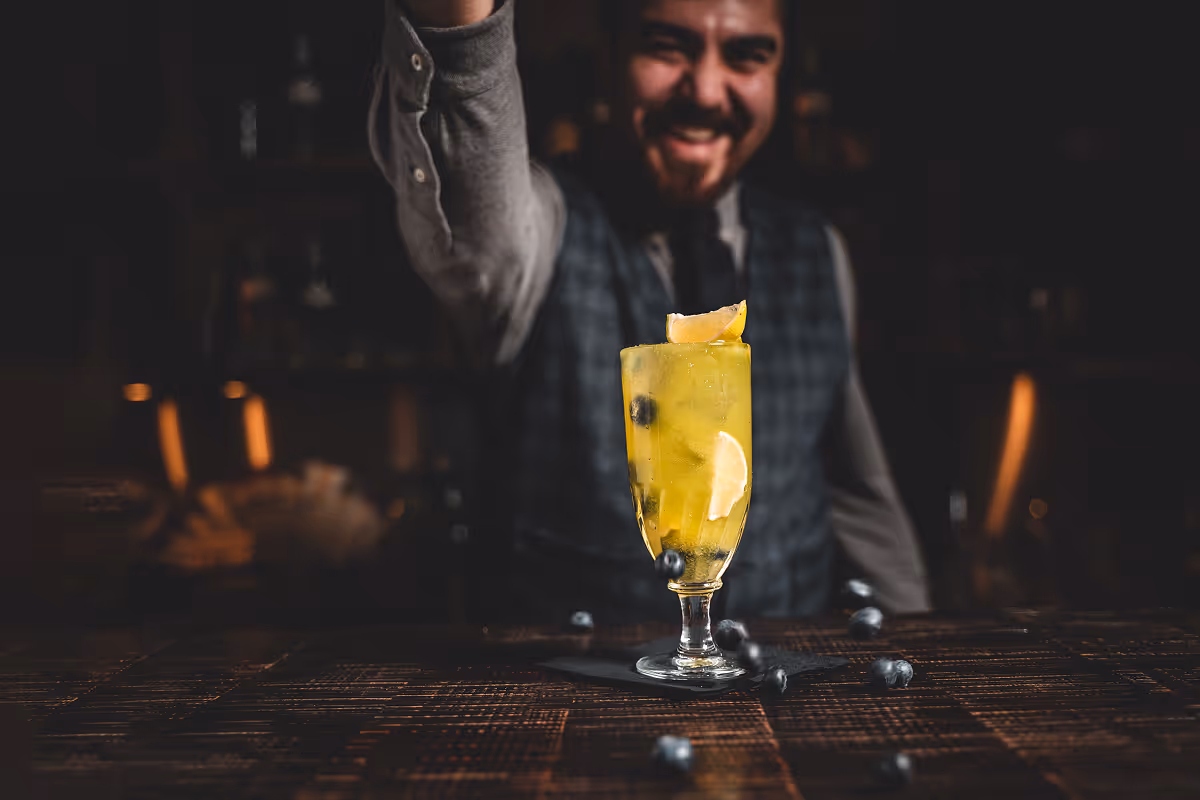 Barkeeper serviert ein gelbes Cocktailgetränk mit Zitronenscheiben und Blaubeeren in einem hohen Glas.