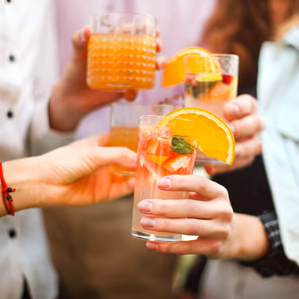 Vier Hände halten Cocktailgläser mit orangefarbenen Getränken und Garnitur, die gemeinsam anstoßen.