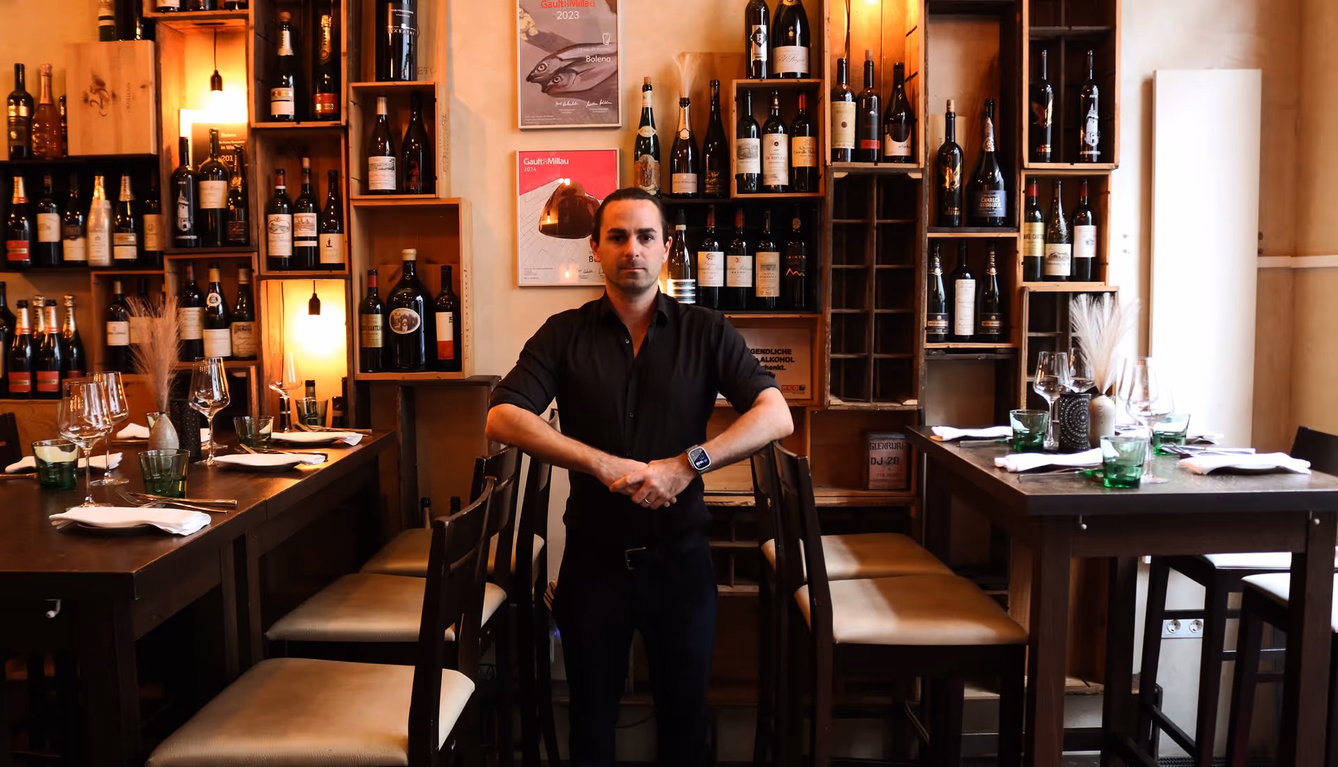 Mann in schwarzem Hemd steht zwischen zwei gedeckten Holztischen mit Weinflaschenregalen an der Wand im Hintergrund.