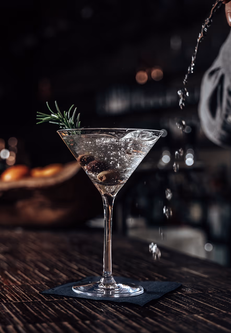 Ein Martini-Cocktail mit Oliven und Rosmarinzweig, daneben werden Tropfen eingegossen.