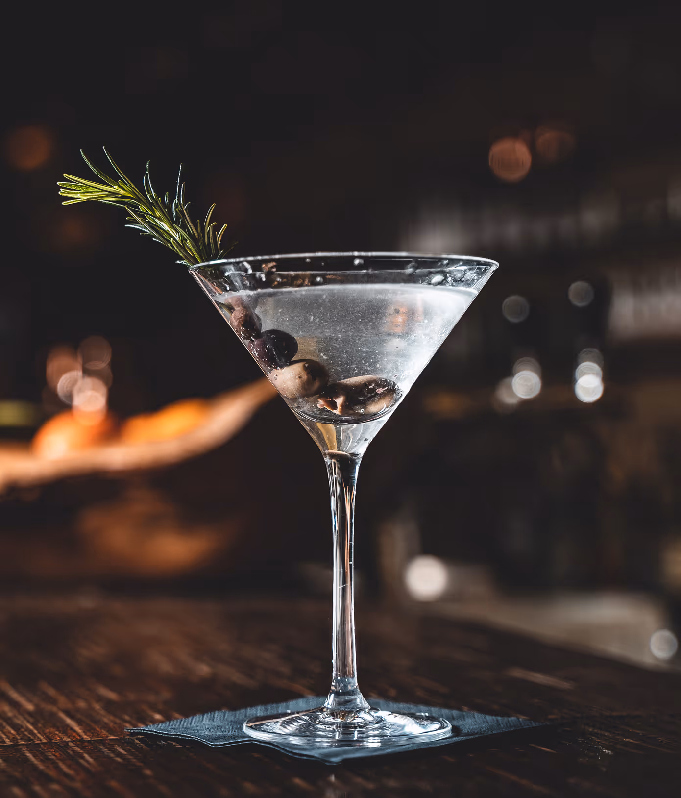 Ein Martini-Cocktail in einem stilvollen Glas mit Oliven und einem Zweig Rosmarin auf einem dunklen Untergrund.