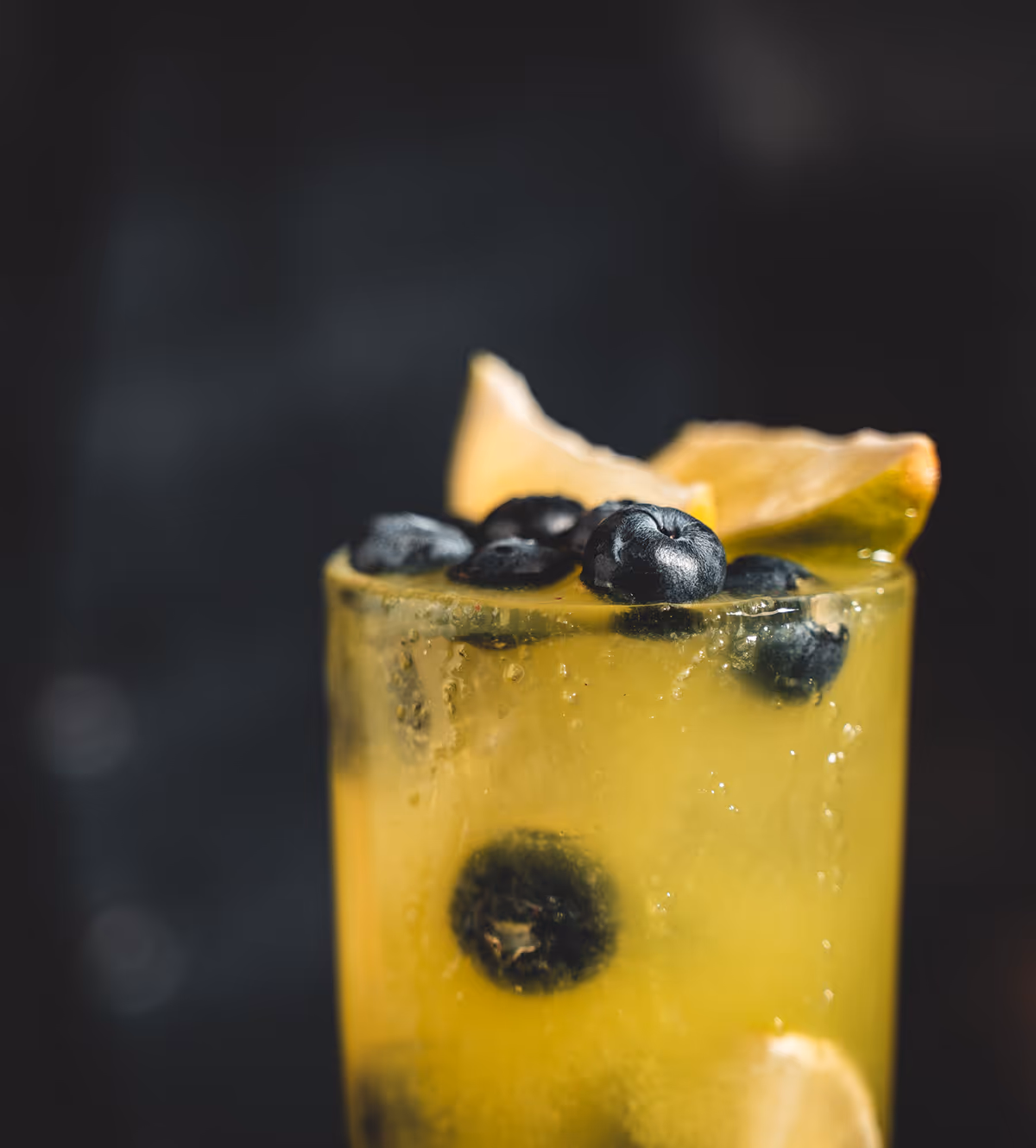 Gelbes Getränk in Glas mit Eiswürfeln, Blaubeeren und Zitronenscheibe vor dunklem Hintergrund.