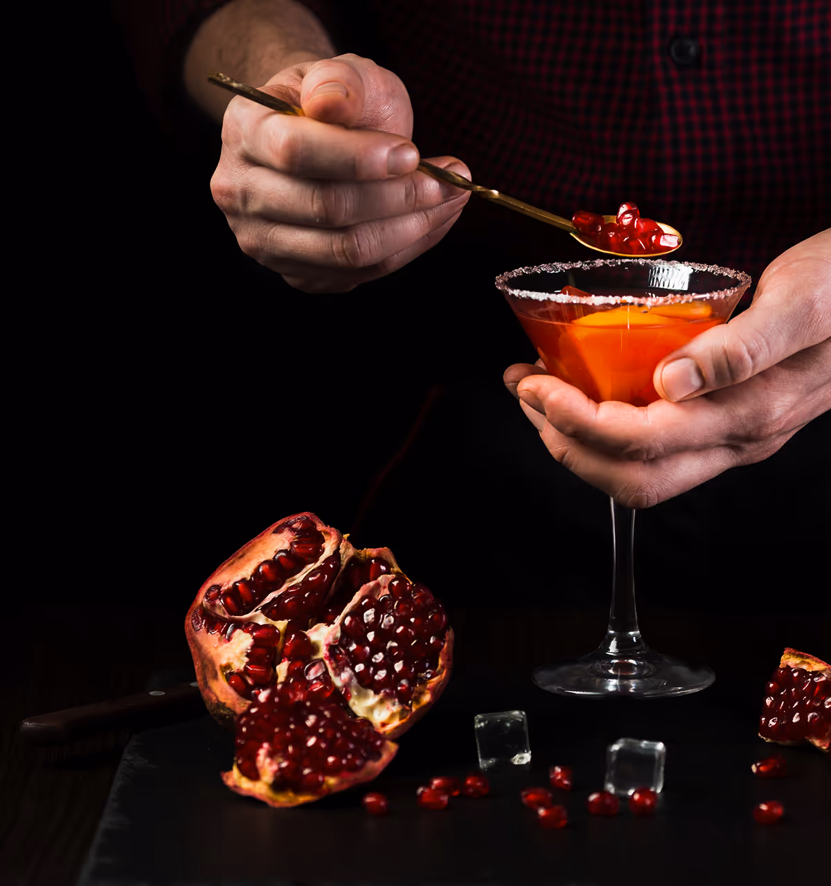 Person fügt mit einem goldenen Löffel Granatapfelkerne in ein Cocktailglas mit Zuckerrand und Orangenscheibe ein, vor schwarzem Hintergrund.