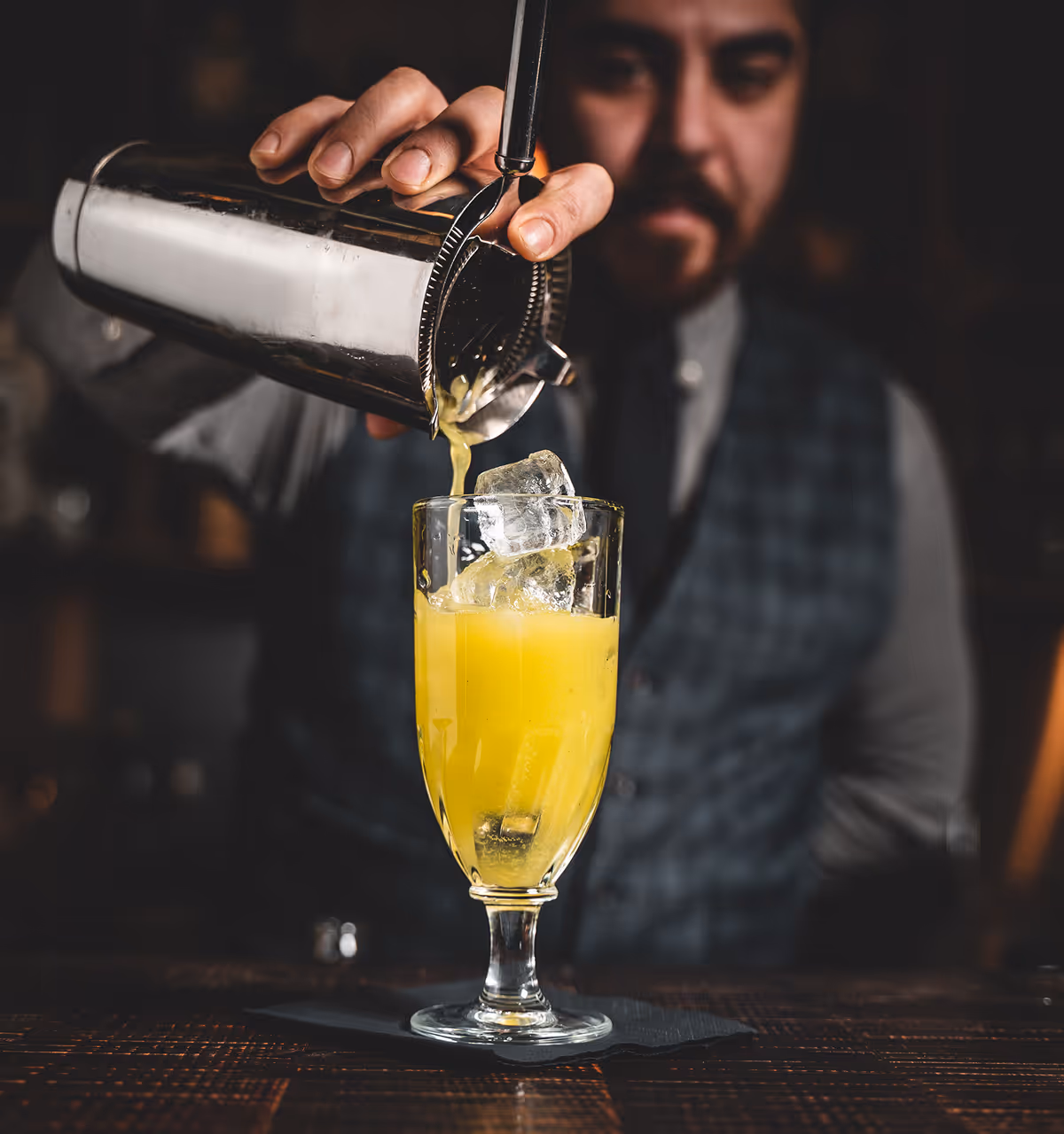 Ein Barkeeper gießt einen gelben Cocktail mit Eiswürfeln aus einem Shaker in ein Glas.
