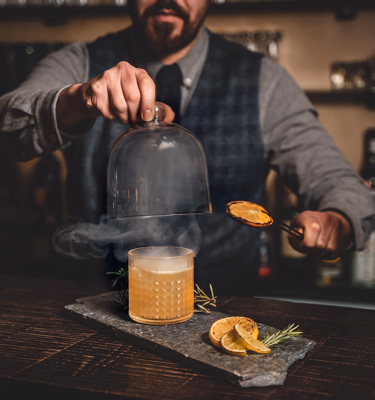 Barkeeper hebt eine Glasglocke von einem rauchenden Cocktailglas auf einem Steinbrett mit getrockneten Zitrusscheiben und Rosmarin.
