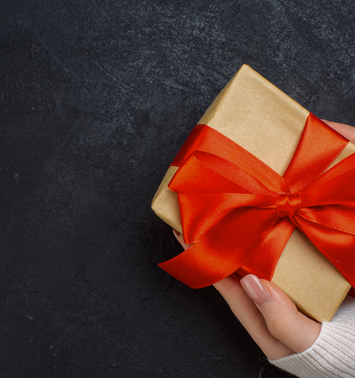 Hand hält ein Geschenk mit goldener Verpackung und roter Schleife vor schwarzem Hintergrund.