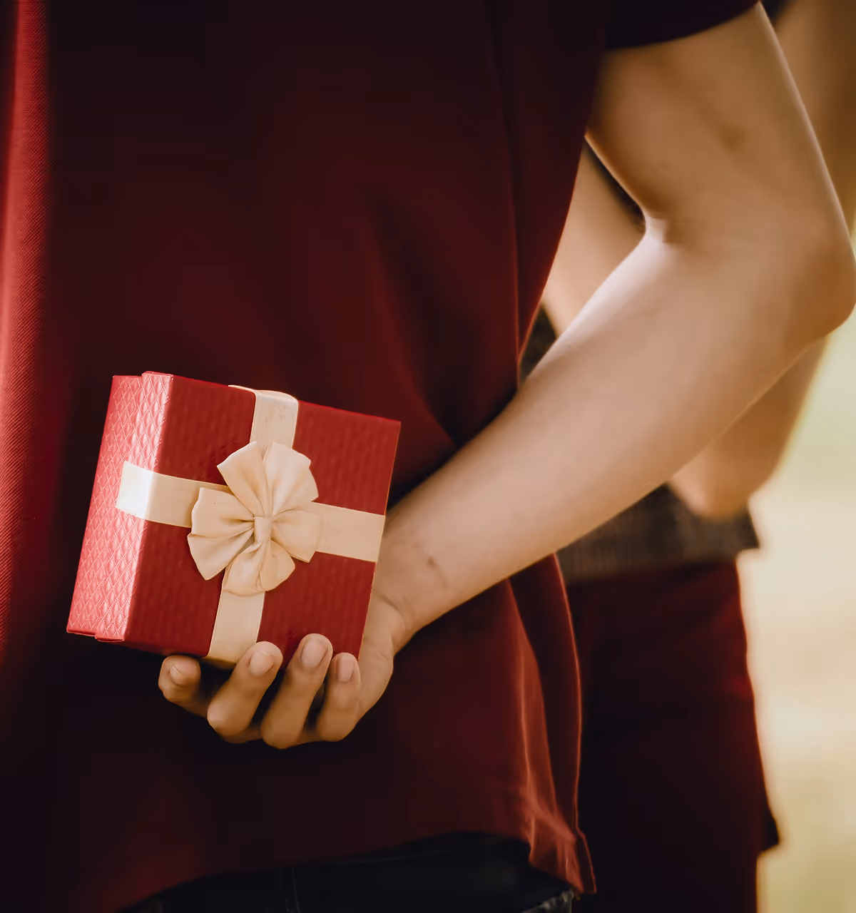 Person hält heimlich ein rot verpacktes Geschenk mit goldener Schleife hinter dem Rücken.