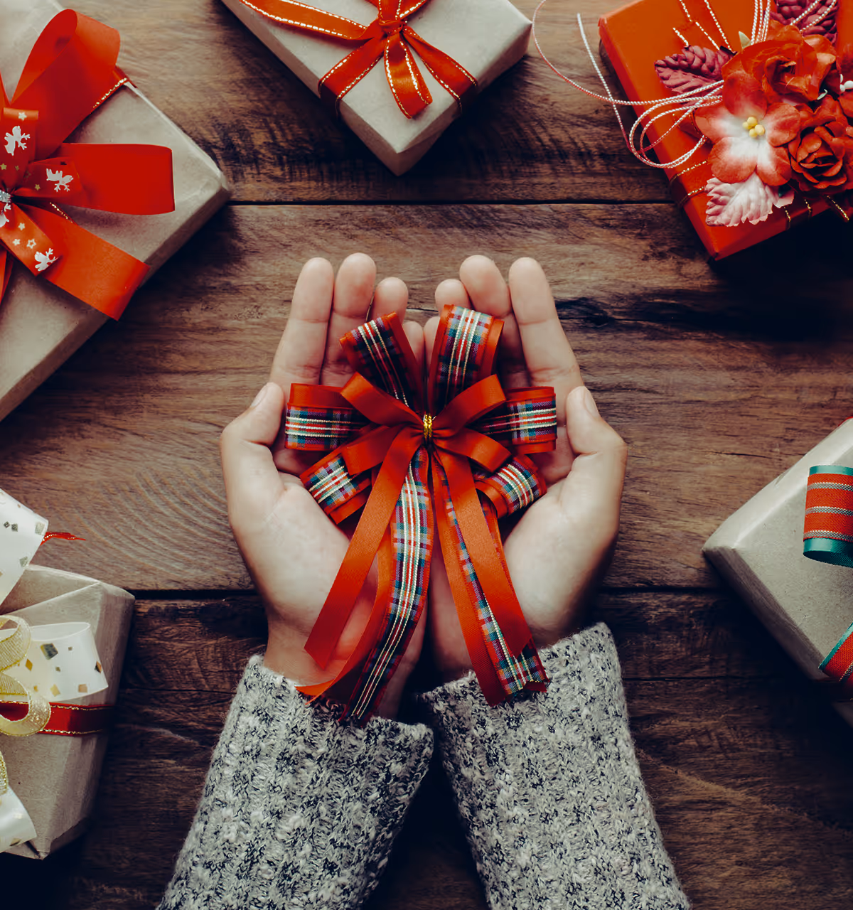 Hände halten eine rote Schleife mit kariertem Muster, umgeben von Geschenkboxen mit roten Schleifen auf einem Holztisch.