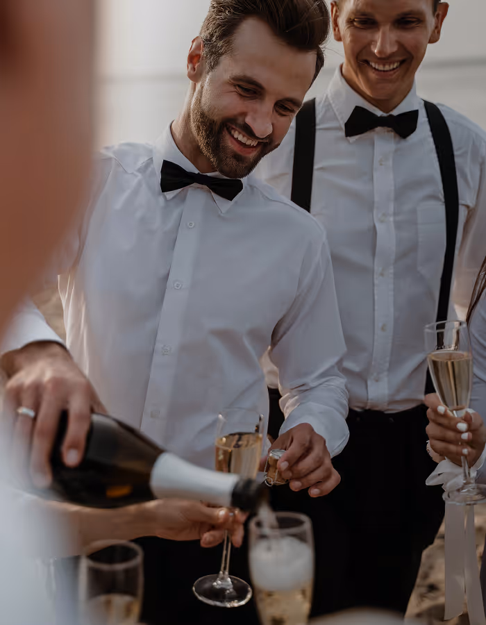 Zwei Männer in weißen Hemden und schwarzen Fliegen lachen, während Champagner in Gläser eingeschenkt wird.