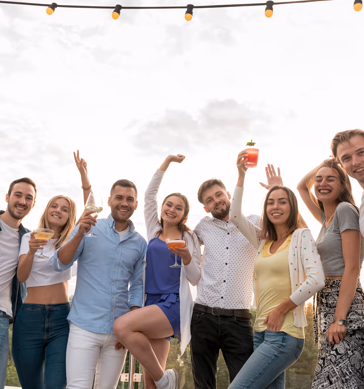 Sieben junge Erwachsene feiern draußen bei Sonnenuntergang mit Getränken in der Hand und Lichterkette darüber.