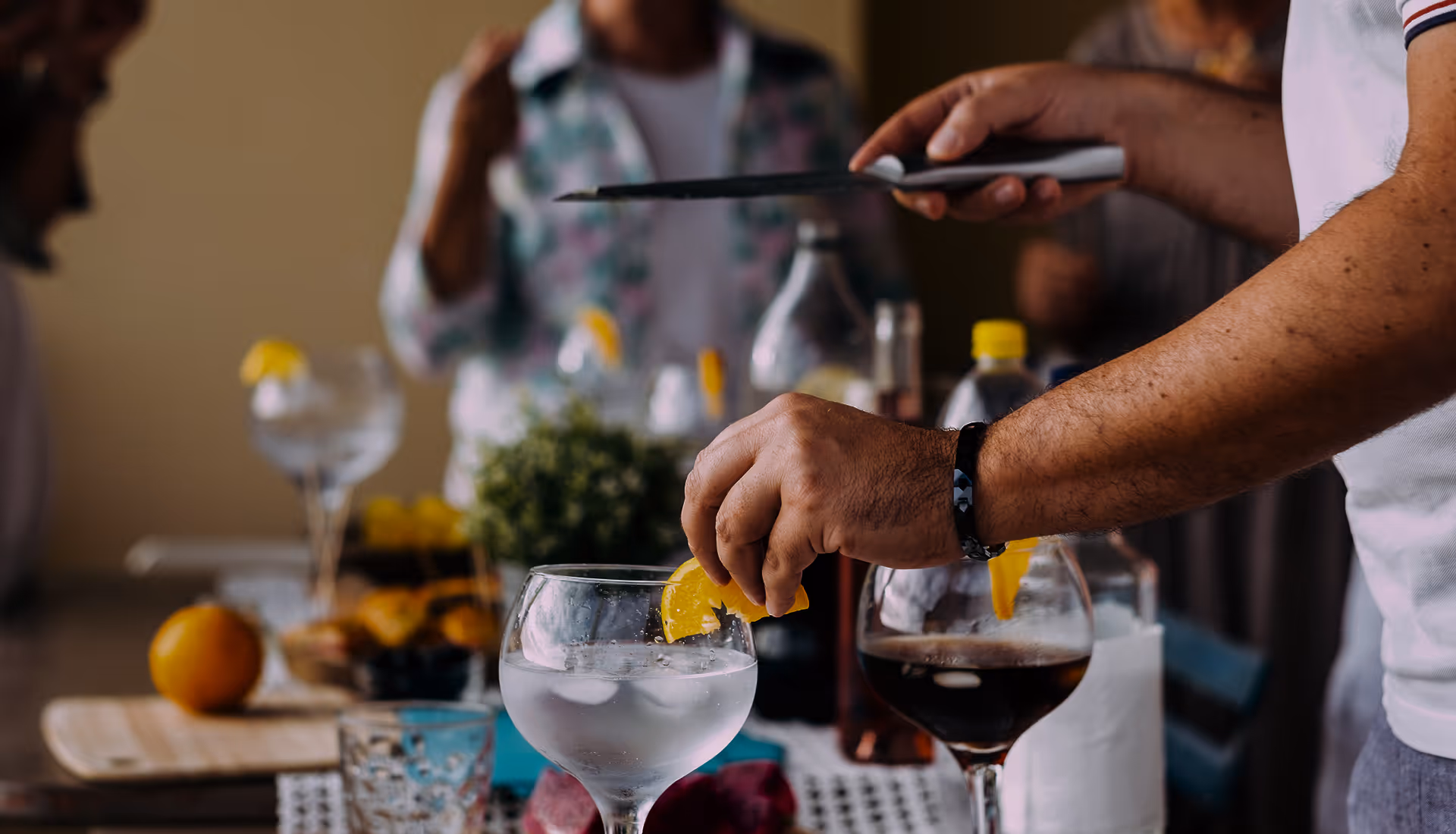 Eine Hand legt eine Orangenscheibe an den Rand eines mit Eis gefüllten Getränks in einem Glas, im Hintergrund Menschen und weitere Getränke.