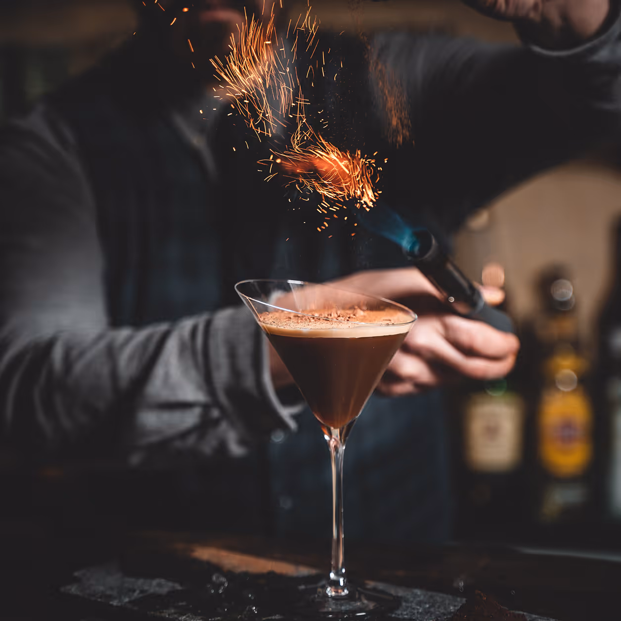 Eine Person bestreut mit einem Brenner Funken auf einen Cocktail in einem Martiniglas.