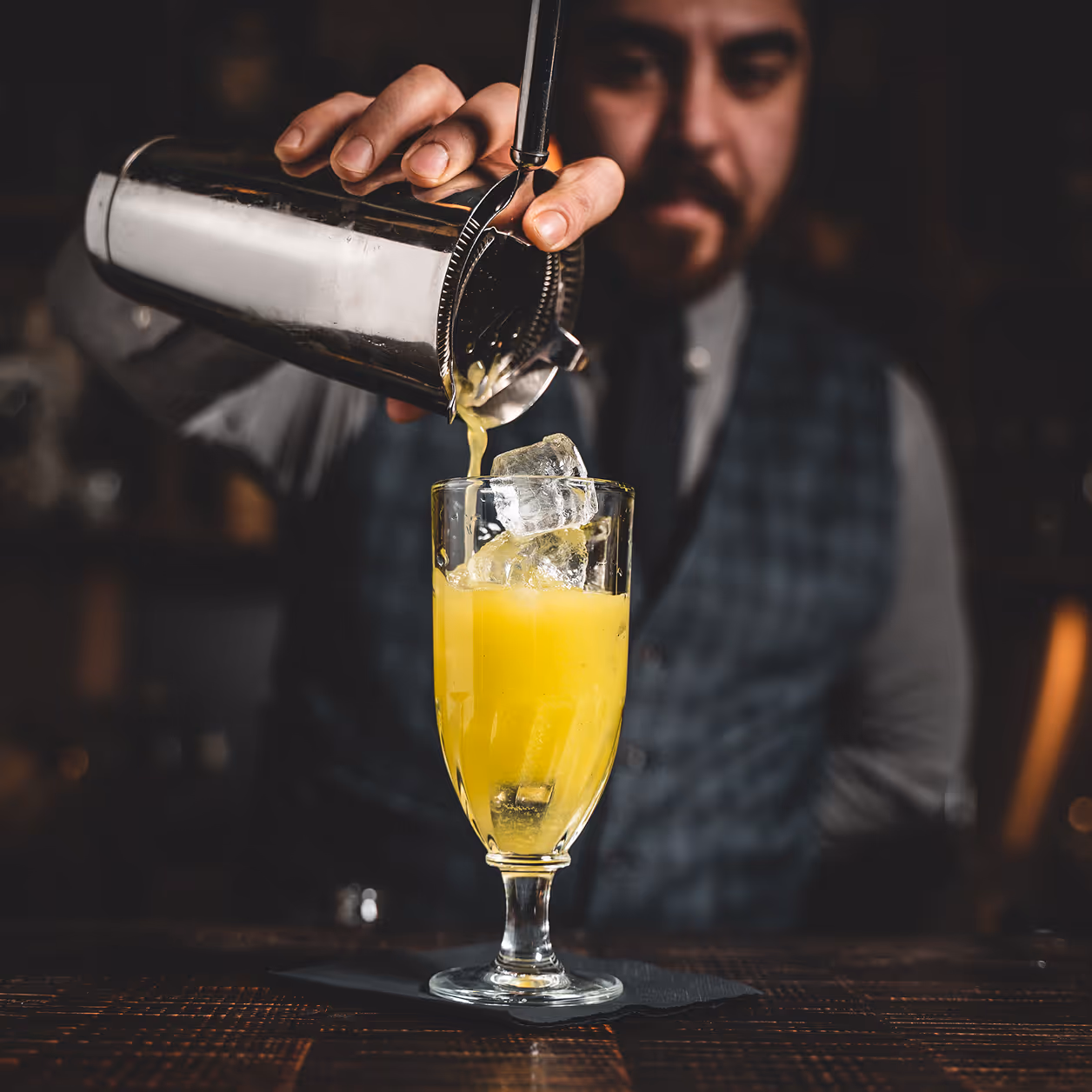 Barkeeper gießt gelben Cocktail mit Eiswürfeln aus einem Shaker in ein Glas.