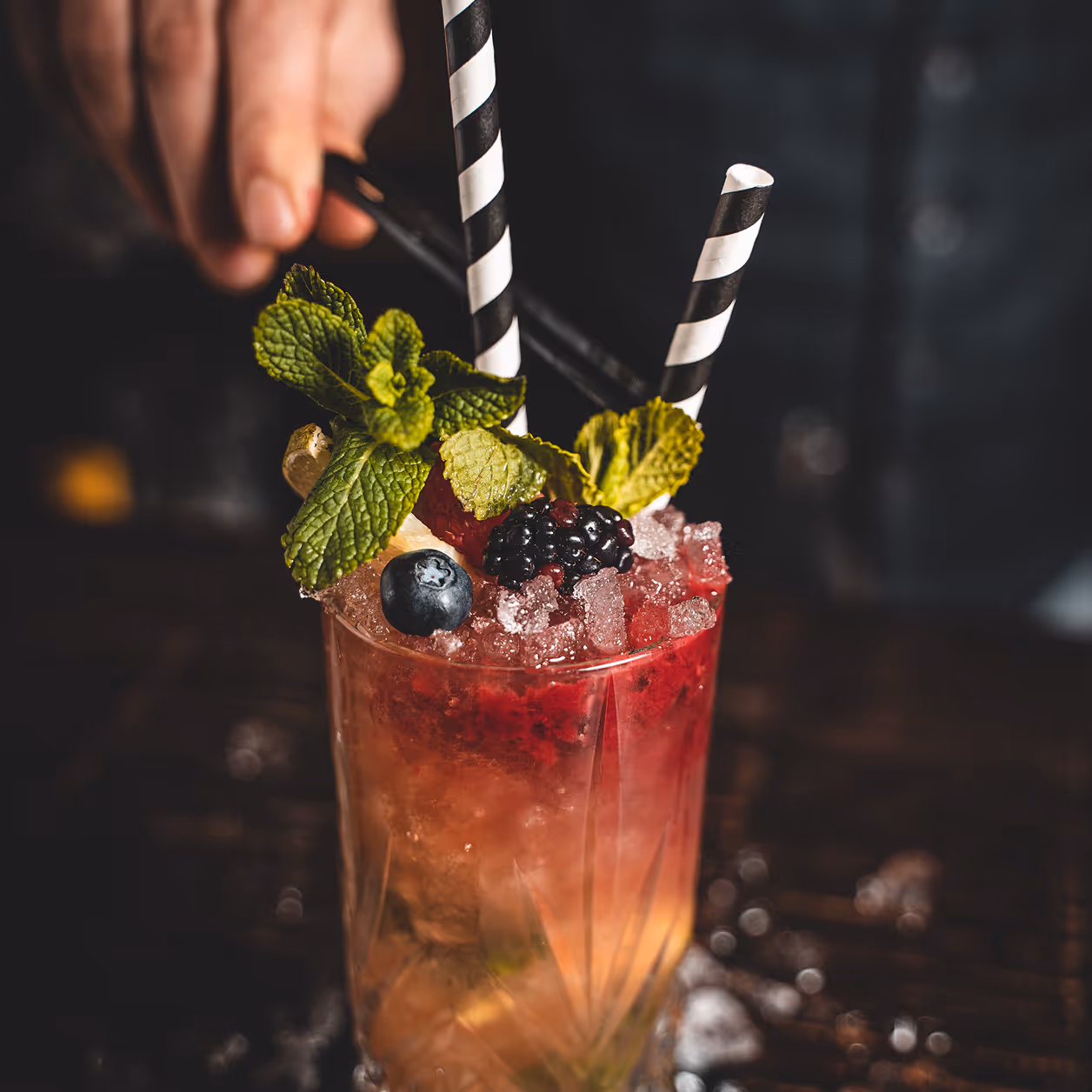 Nahaufnahme eines roten Cocktails mit zerstoßenem Eis, Beeren, Minzblättern und zwei schwarz-weißen Strohhalmen.