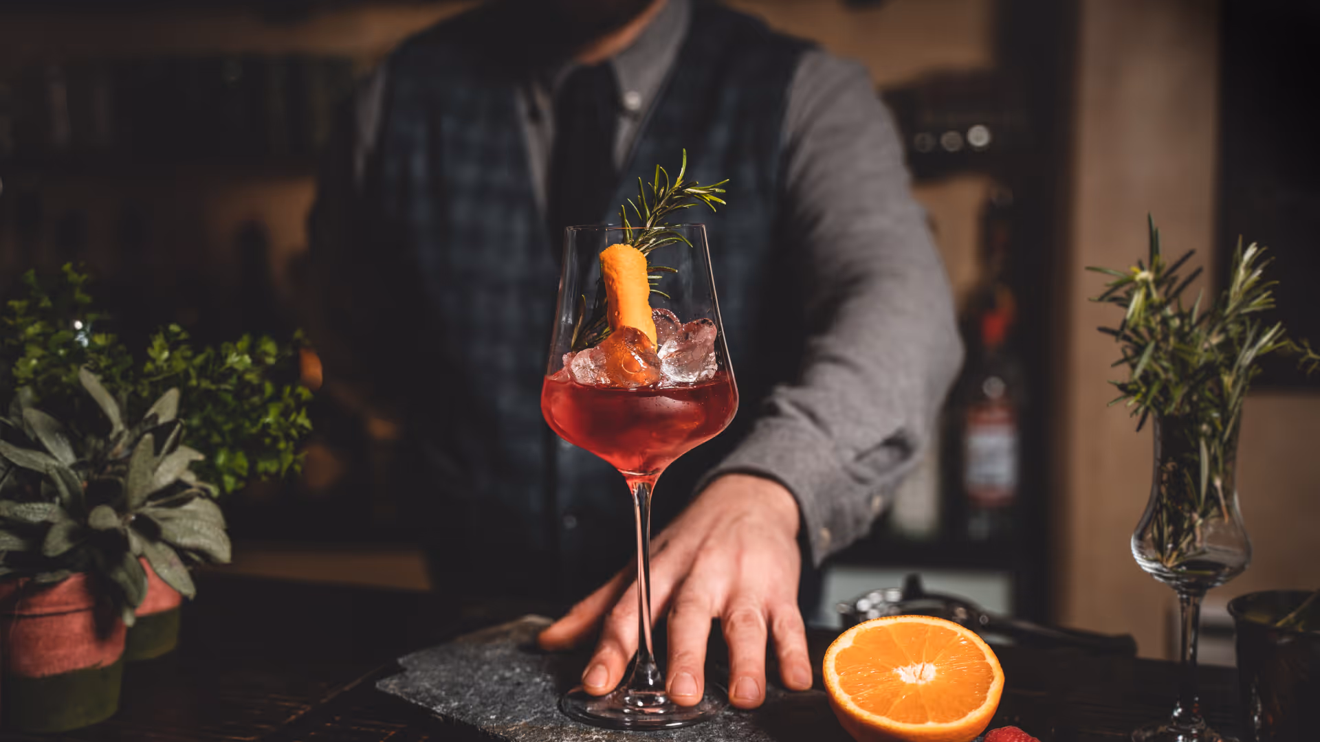 Ein Barkeeper serviert einen roten Cocktail im Weinglas mit Eis, einer Karotte und einem Rosmarinzweig, daneben liegt eine halbe Orange.