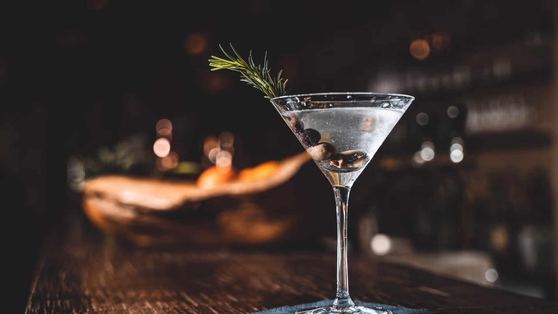 Martini-Glas mit Klarem Getränk, gefüllt mit Oliven und garniert mit einem Rosmarinzweig auf einem Holztisch.