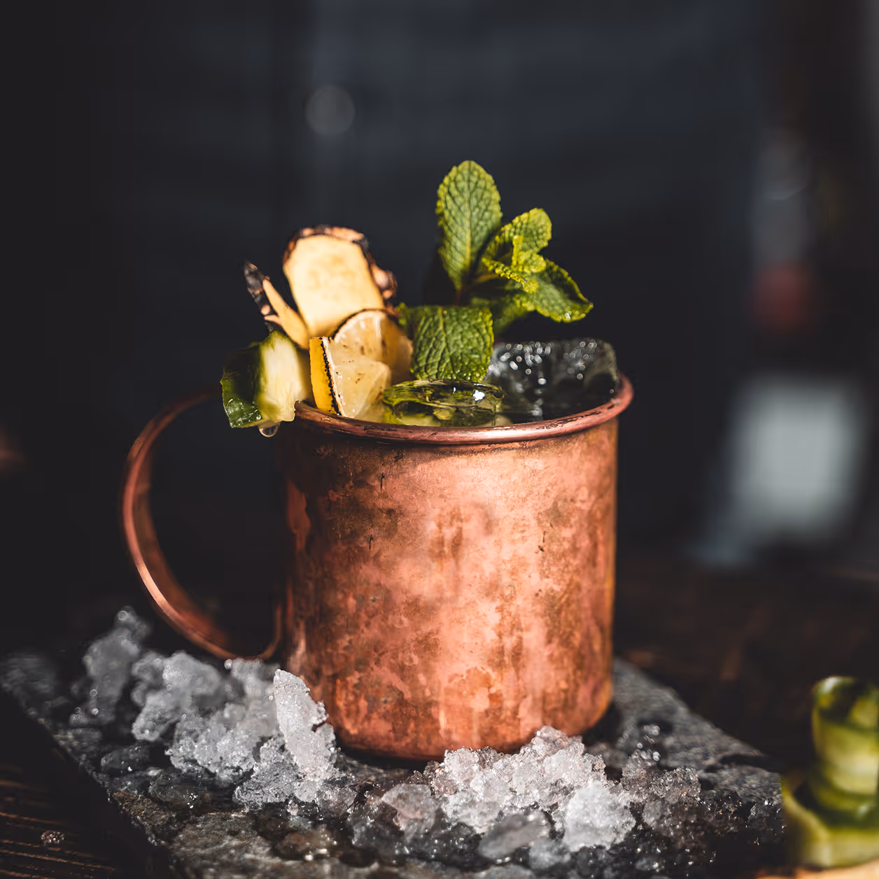 Kupfertasse mit Eiswürfeln, Gurken-, Zitronen- und Ingwerscheiben sowie frischer Minze auf einer mit zerstoßenem Eis bedeckten Oberfläche.