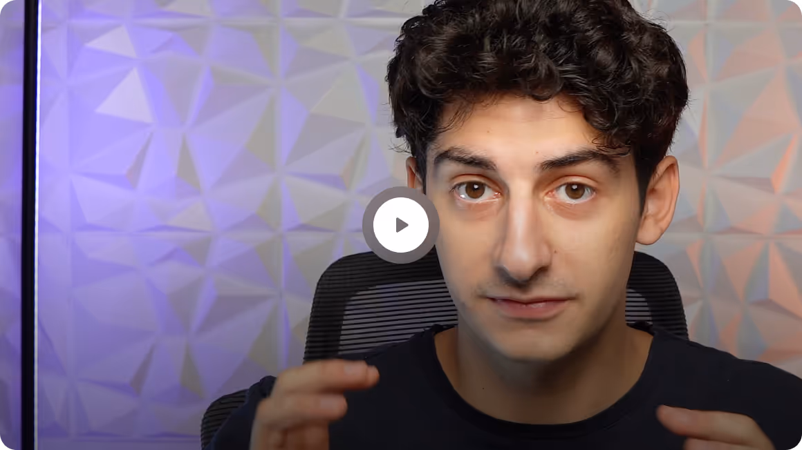 Young man with curly dark hair explaining something in front of a geometric patterned wall with soft purple and orange lighting.