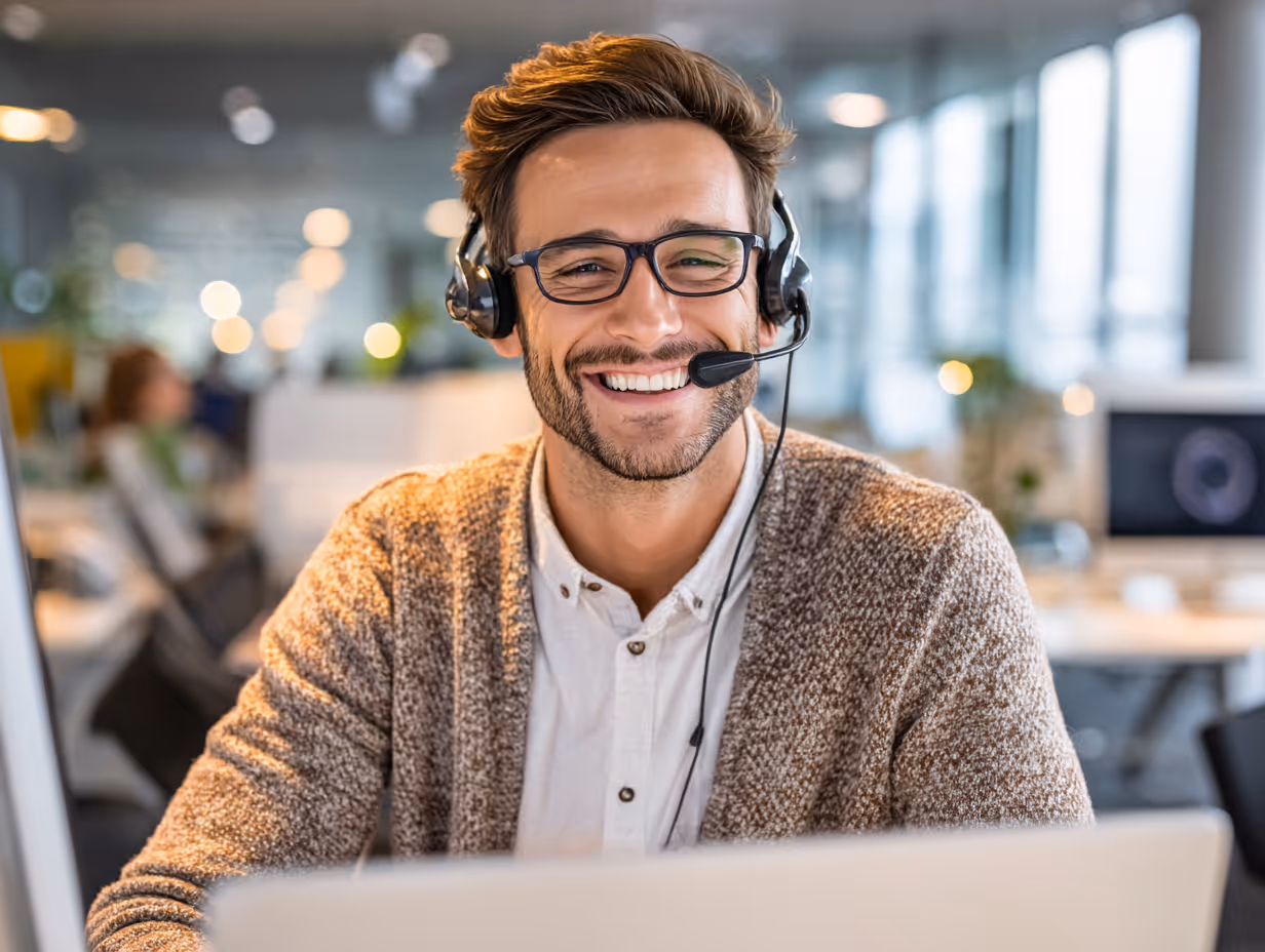 Smiling man wearing glasses and headset working on a laptop in a modern office.