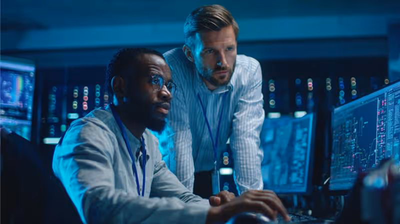 Two men focused on computer screens displaying data in a high-tech control room.