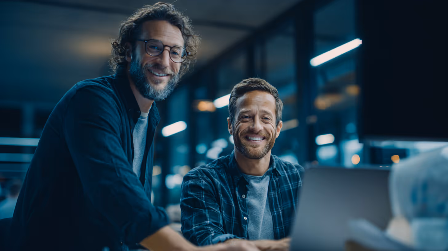 Two men smiling while working together on a laptop in a modern office with large windows at night.