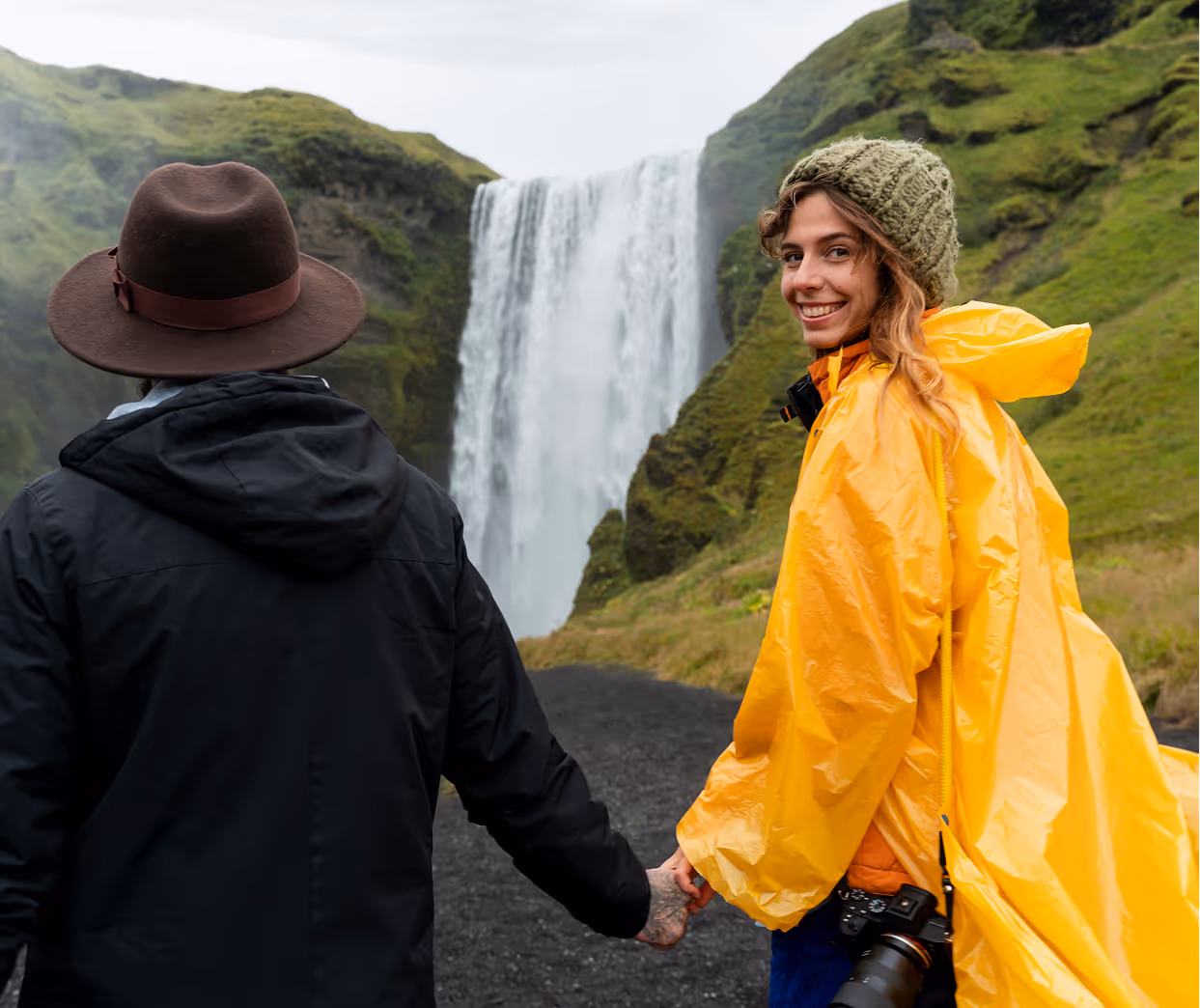Couple holding hands with a woman in a yellow raincoat smiling at the camera near a large waterfall surrounded by green hills.