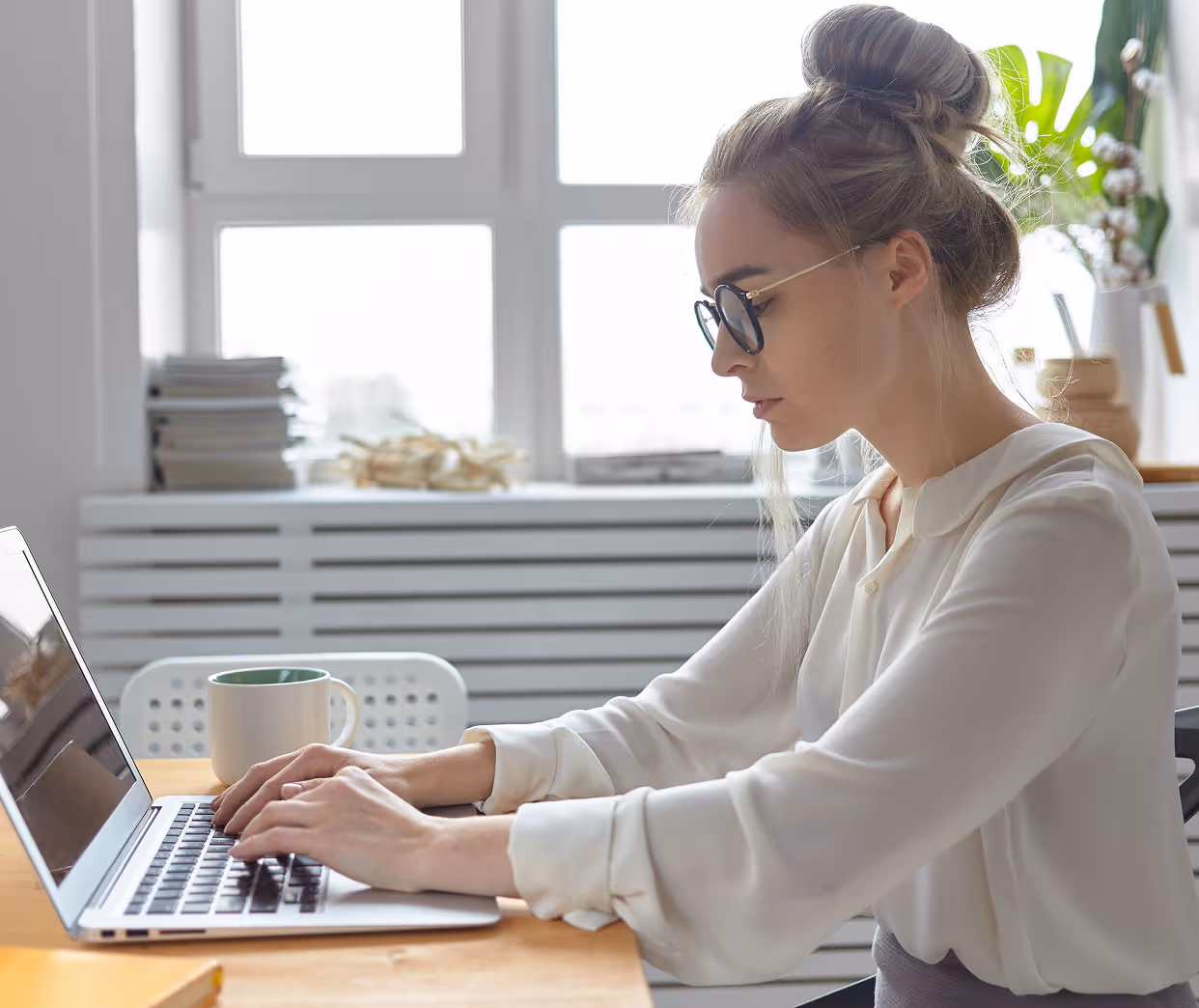 Woman with glasses and hair in a bun typing on a laptop at a wooden desk with a coffee mug nearby.
