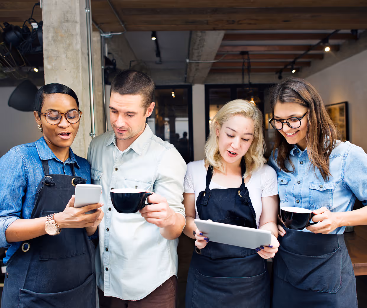 Four cafe staff wearing aprons looking at a phone and tablet, holding coffee cups and smiling.