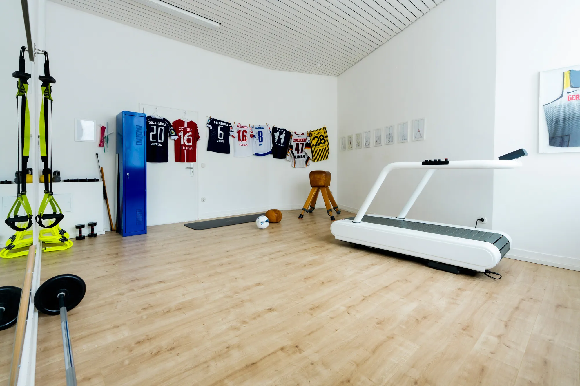Weitwinkelansicht eines modernen Trainingsraums mit Laufband, Trainingsgeräten, Sporttrikots an der Wand und hellem Holzboden