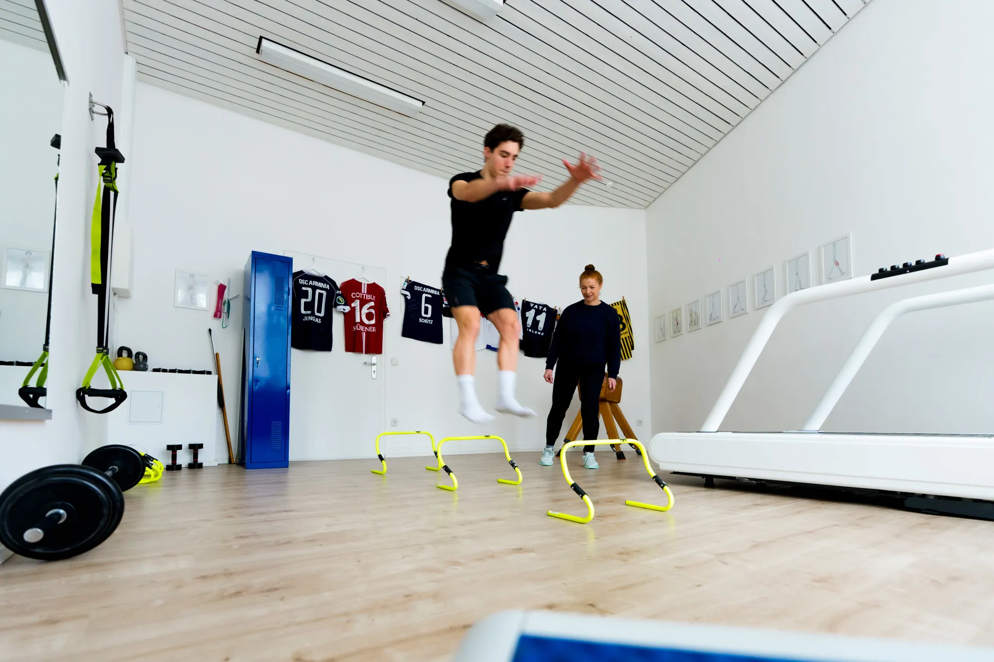 Ein junger Sportler springt über gelbe Trainingshürden, während Jessica Mathiak im Hintergrund beobachtet, moderner Trainingsraum mit Sporttrikots an der Wand