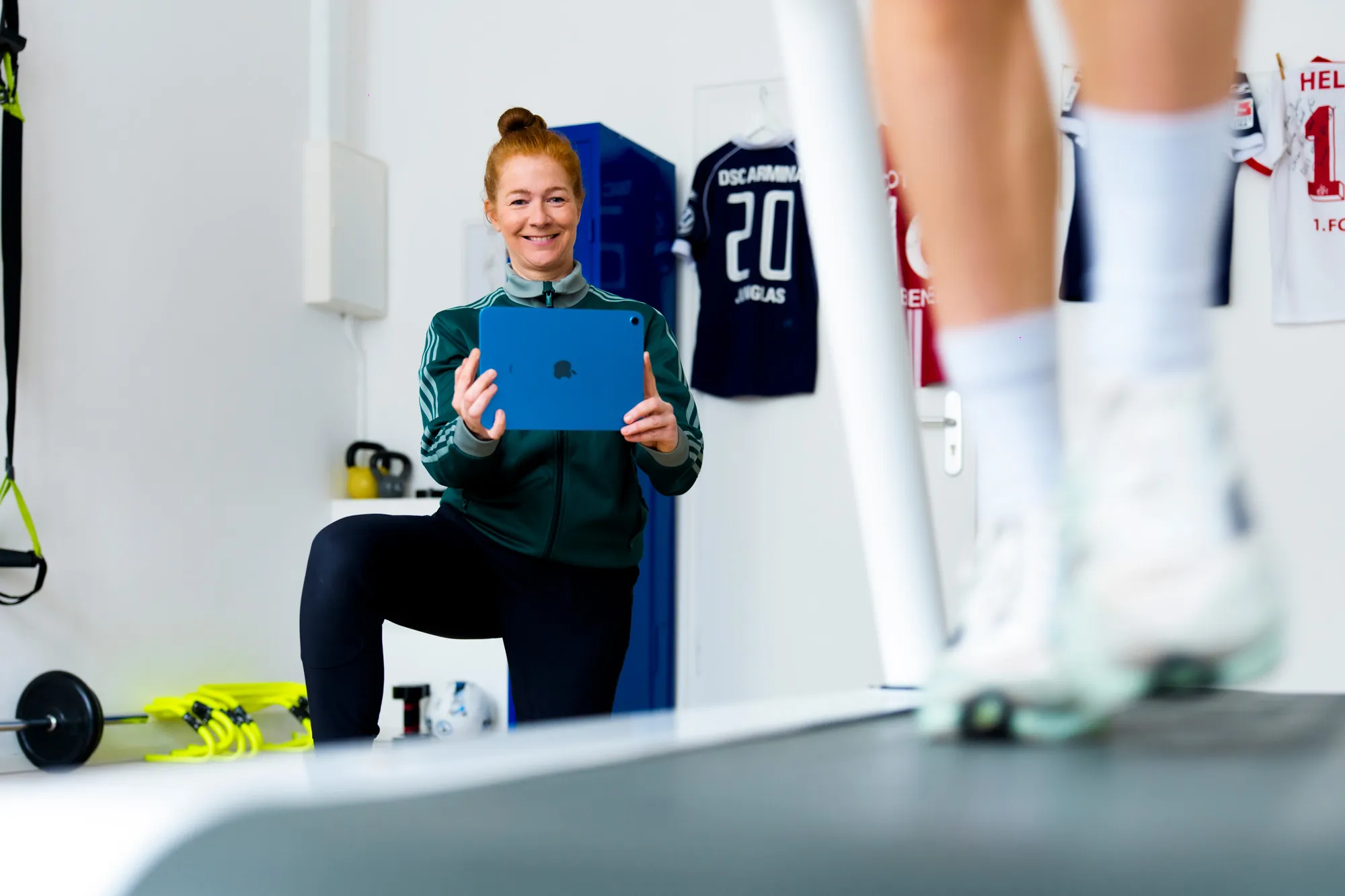 Jessica Mathiak in grünem Trainingsanzug hält ein iPad, während sie eine Person im Training beobachtet und eine Gang- und Laufanalyse durchführt, Trainingsraum mit Sporttrikots im Hintergrund