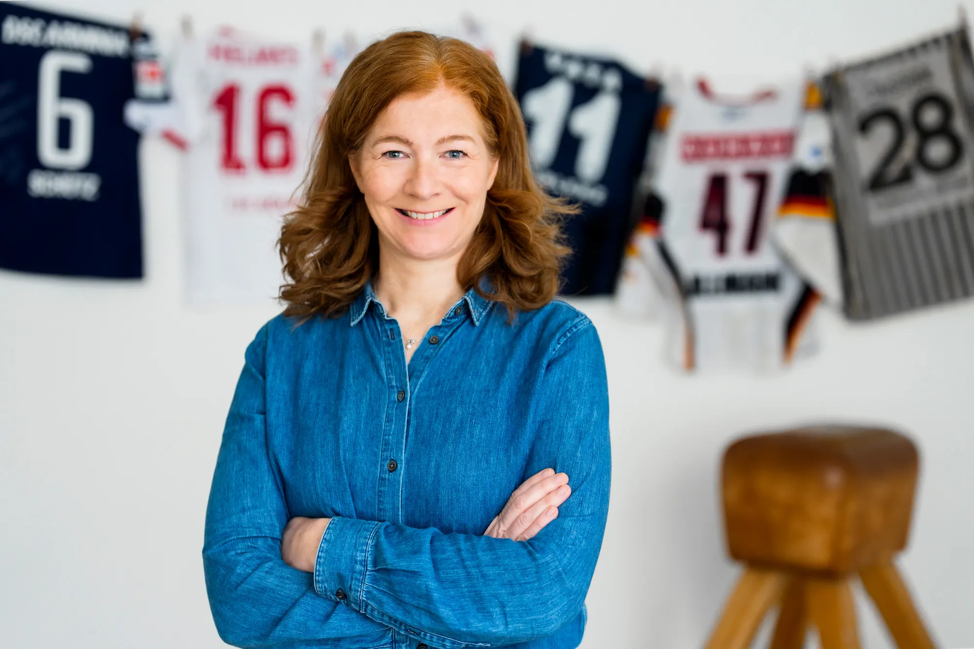 Jessica Mathiak in blauem Jeanshemd mit verschränkten Armen, stehend in einem hellen Trainingsraum mit Sporttrikots im Hintergrund