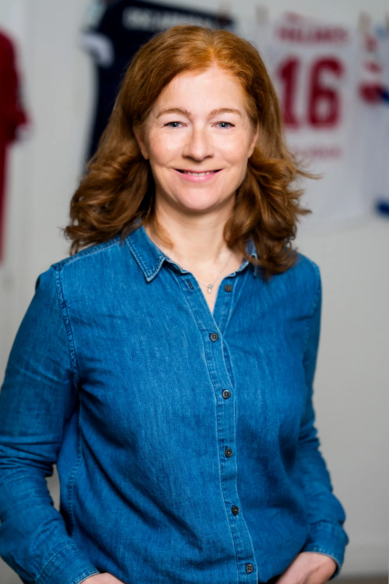 Portrait von Jessica Mathiak mit roten Haaren und blauem Jeanshemd, lächelnd vor einem Hintergrund mit Sporttrikots